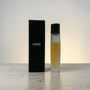 Könnte beinhalten: Ein Parfümflaschen- und Box-Set. Die Flasche hat eine klare, rechteckige Form mit schwarzem Verschluss und enthält eine hell bernsteinfarbene Flüssigkeit. Die Box ist schwarz mit dem weißen Aufdruck "mania".