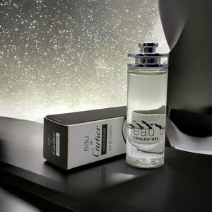 Puede incluir: Un frasco de perfume de vidrio transparente con una tapa plateada y las palabras "eau de concentree" impresas en la parte delantera. Una caja en blanco y negro con las palabras "Cartier" y "eau de concentree" está al lado del frasco. El fondo es una superficie brillante.