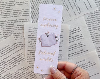 May include: A white bookmark with a pink and purple illustration of a book with flowers and mountains. The bookmark has the text "forever exploring fictional worlds" in pink and purple lettering.