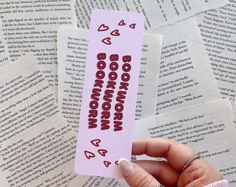 May include: A pink and white bookmark with the words "BOOKWORM" in bold, red letters. The bookmark is decorated with small pink hearts.
