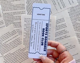 May include: A blue and white bookmark with the text "Sports Romance Book Club" and "Admit One". The bookmark is held in a hand with a silver ring on the finger.