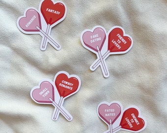 May include: Five pairs of heart-shaped lollipop stickers with different romantic phrases. The phrases are "Romance", "Fantasy", "Small Town", "Cowboy Romance", "Fake Dating", "Friends to Lovers", "Fated Mates", and "Enemies to Lovers".