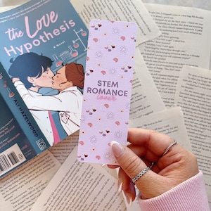 May include: A purple and white bookmark with a pattern of hearts, brains, and atoms. The bookmark has the text "STEM Romance Lover" in black.