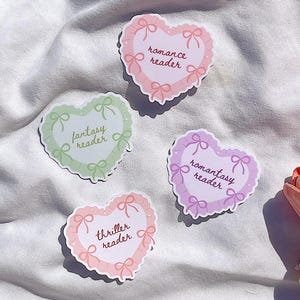 Puede incluir: Cuatro pegatinas en forma de corazón en tonos pastel rosa, verde y morado, cada una con un diseño de lazo. Las pegatinas presentan las frases "romance reader", "fantasy reader", "thriller reader" y "romantasy reader".