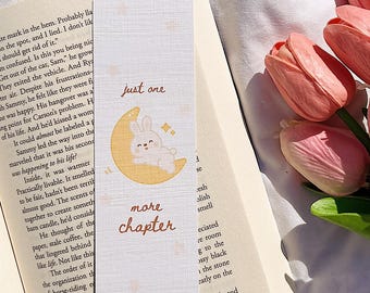May include: A bookmark with a cute bunny on a crescent moon, the text "just one more chapter" is written on the bookmark. The bookmark is placed in an open book with pink tulips in the background.