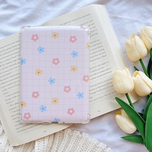 May include: A light pink tablet case featuring a grid pattern and colourful flower designs. The case is on an open book, beside a bouquet of white tulips and a white knitted blanket. The book has white pages.