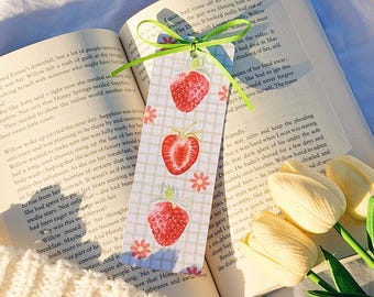 May include: A handmade bookmark featuring three red strawberries with green leaves and pink flowers on a checkered background. The bookmark is placed in an open book with text, next to white tulips.