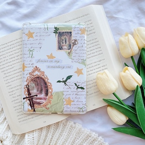 May include: Transparent phone case with a collage design. It features a mirror, a sword, and the text "forever in my romantasy era." The case has gold star accents and a small image of a doorway. The case is on an open book.