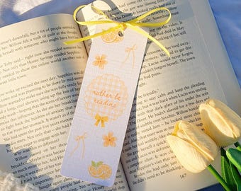 May include: A decorative bookmark with a yellow ribbon, featuring a repeating pattern of orange slices, flowers, and bows. The bookmark has the text "rather be reading" in a playful font. The bookmark is resting on an open book.