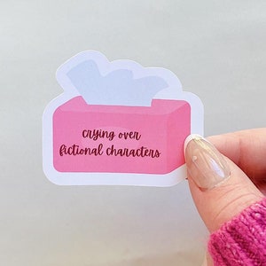Puede incluir: Una pegatina rosa con forma de caja de pañuelos con el texto "crying over fictional characters". La pegatina tiene un borde blanco y un pañuelo blanco que sobresale de la parte superior. La pegatina la sostiene una persona que lleva un jersey rosa.