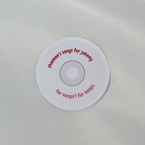 May include: White round sticker with pink text that reads "Shannon's songs for Johnny for keeps? for keeps."