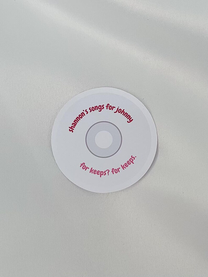 May include: White round sticker with pink text that reads "Shannon's songs for Johnny for keeps? for keeps."