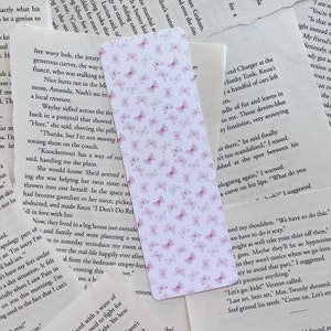 Op de afbeelding: Een roze en witte bladwijzer met een delicaat vlinderpatroon. De bladwijzer is gemaakt van papier en heeft een subtiele textuur. De vlinders zijn verspreid over de bladwijzer, wat een speels en elegant ontwerp cre&euml;ert.