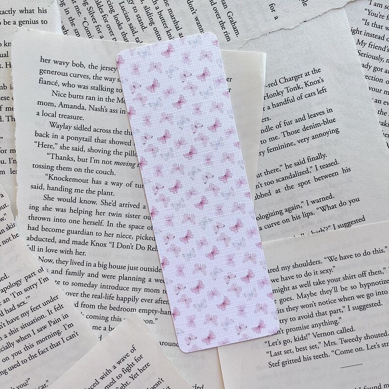 Op de afbeelding: Een roze en witte bladwijzer met een delicaat vlinderpatroon. De bladwijzer is gemaakt van papier en heeft een subtiele textuur. De vlinders zijn verspreid over de bladwijzer, wat een speels en elegant ontwerp cre&euml;ert.