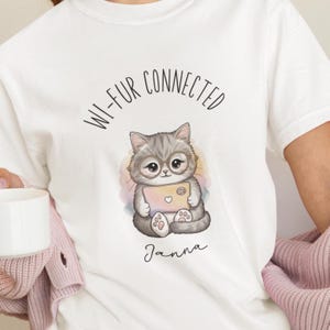 May include: White t-shirt with a graphic of a gray cat wearing glasses and holding a laptop. The text "WI-FUR CONNECTED" arches above the cat, with the name "Janna" below. The cat is surrounded by pastel colors.