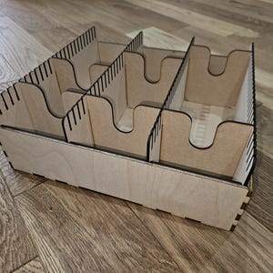May include: A light-coloured wooden organiser with multiple compartments. The organiser has a rectangular base and is divided into sections with curved cutouts. The dividers have vertical slots. The item is designed for storage and organisation.