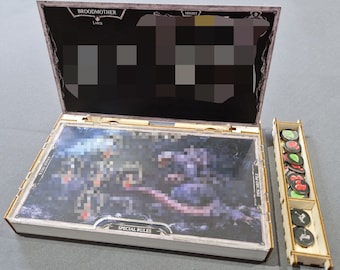 May include: A wooden box with a hinged lid, containing a card game with a fantasy illustration. The box has a label that reads "Broodmother" and "Special Rules". The box is open and the card game is visible. There is a separate wooden tray with compartments containing small tokens.