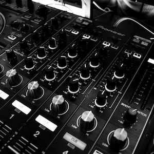 May include: A black DJ mixer with multiple knobs and buttons, including a large knob labeled "CUE" and a smaller knob labeled "BOOT/MONITOR". The mixer has a silver faceplate with the word "Pioneer" printed on it.