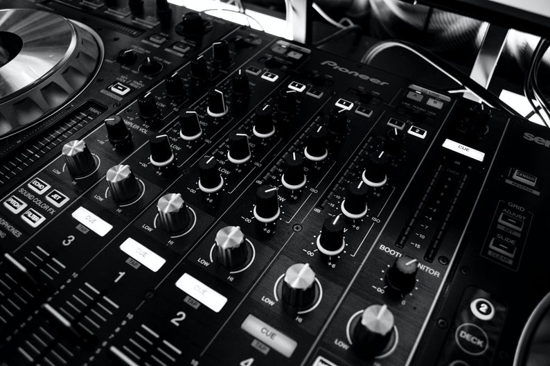 May include: A black DJ mixer with multiple knobs and buttons, including a large knob labeled "CUE" and a smaller knob labeled "BOOT/MONITOR". The mixer has a silver faceplate with the word "Pioneer" printed on it.