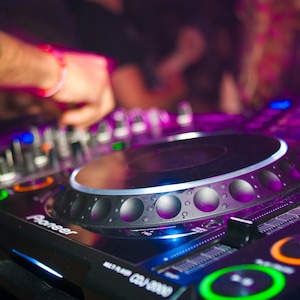 May include: A close-up of a black Pioneer DJ mixer with a silver control knob and a purple light reflecting on the surface. The mixer has a variety of buttons and knobs for controlling music.