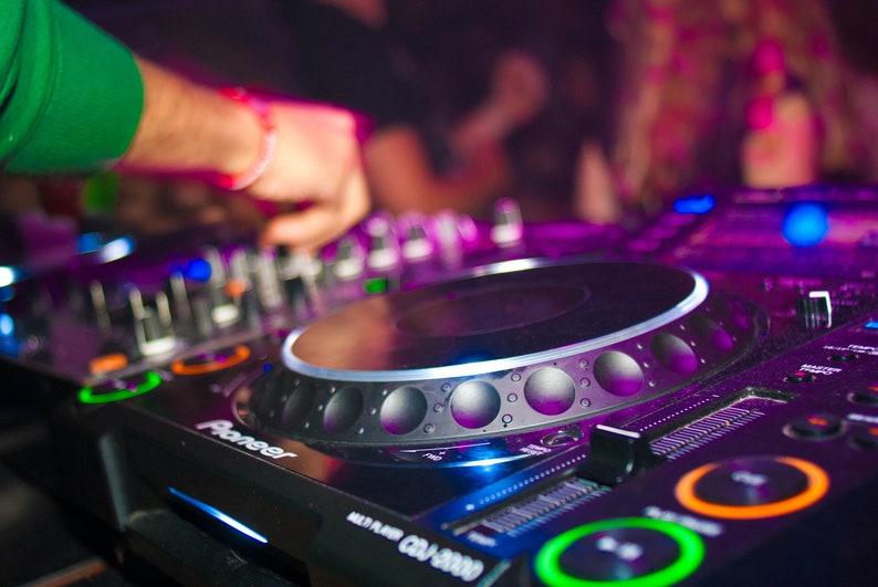 May include: A close-up of a black Pioneer DJ mixer with a silver control knob and a purple light reflecting on the surface. The mixer has a variety of buttons and knobs for controlling music.