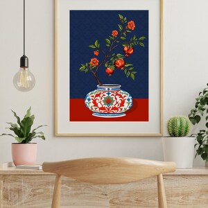 May include: A framed wall art print featuring a decorative ceramic vase with a white base, red floral patterns, and blue accents. A branch with green leaves, orange flowers, and red pomegranate fruits emerges from the vase against a dark blue background.