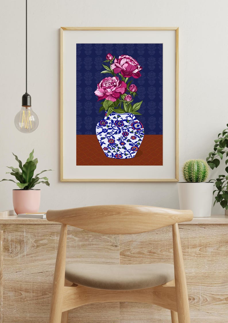 May include: A framed wall art print featuring a white vase with blue and red floral patterns containing vibrant pink peony flowers. The background consists of a dark blue patterned wall section above a solid brown surface, all enclosed in a light wood frame.