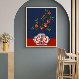 May include: A framed wall art print featuring a decorative white vase with intricate red and blue floral patterns. A branch with green leaves, orange blossoms, and red pomegranate fruits emerges from the vase against a dark blue background.