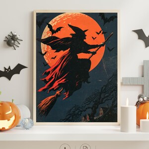 K&ouml;nnte beinhalten: Ein gerahmter Kunstdruck mit der Silhouette einer Hexe auf einem Besen vor einem gro&szlig;en orangefarbenen Vollmond. Die dunkle Szene zeigt fliegende Flederm&auml;use sowie eine Landschaft mit B&auml;umen und H&auml;usern. Der Druck steht auf einem wei&szlig;en Regal.