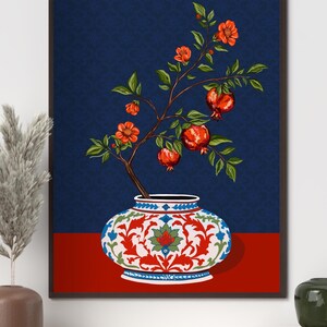 May include: A framed wall art print featuring a white ceramic vase with intricate red, blue, and green floral patterns. A pomegranate branch with green leaves, orange blossoms, and three red pomegranates emerges from the vase against a dark blue background.