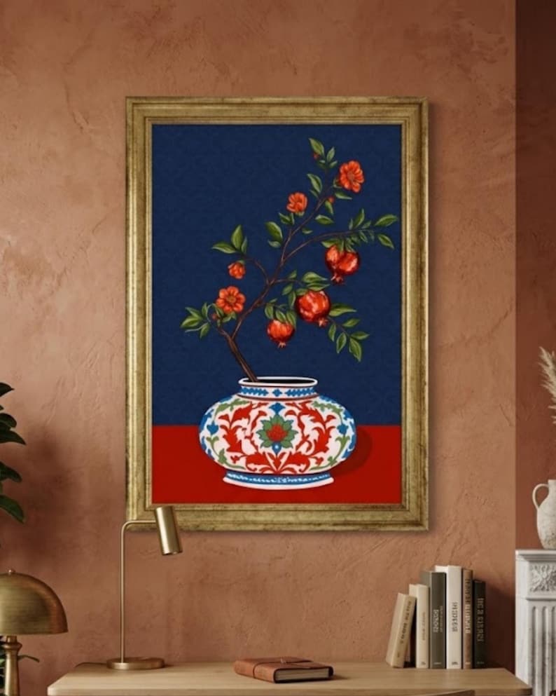 May include: A framed wall art print featuring a decorative vase with a white, red, and blue floral pattern. A branch with green leaves and several red pomegranate fruits extends upward from the vase against a dark blue background.
