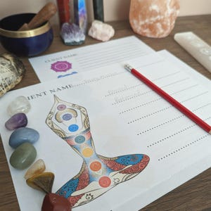 May include: A workspace setup for spiritual practices featuring a printed chart with chakra illustrations, a red pencil, and various gemstones. The chart includes the words "Client Name" and "Blessed, Balanced". Other items include a singing bowl and crystals.