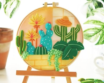 May include: A felt art piece featuring a cactus garden scene. The artwork is displayed in a round embroidery hoop and mounted on a small wooden easel. The scene includes various cacti, flowers, and a straw hat, all in vibrant colors.