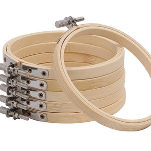 May include: A stack of oval-shaped wooden embroidery hoops with silver-colored adjustable screws. The hoops are made of light-colored wood and are designed for holding fabric taut during embroidery or cross-stitch projects.