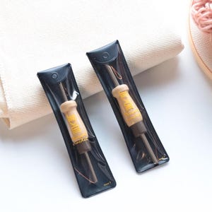 Könnte beinhalten: Zwei Punch-Needle-Werkzeuge mit Holzgriffen und gelbem Text werden in schwarzen Kunststoffhüllen präsentiert. Ein weißer Stoff und ein hölzerner Stickrahmen mit rosa Garn befinden sich im Hintergrund. Die Werkzeuge sind für Stickerei und Handarbeiten.