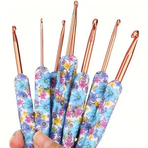 Pu&ograve; includere: Un set di uncinetti con steli in metallo color rame e manici con motivi floreali blu, rosa e bianchi. Gli uncinetti sono tenuti in una mano, mostrando diverse dimensioni e stili.
