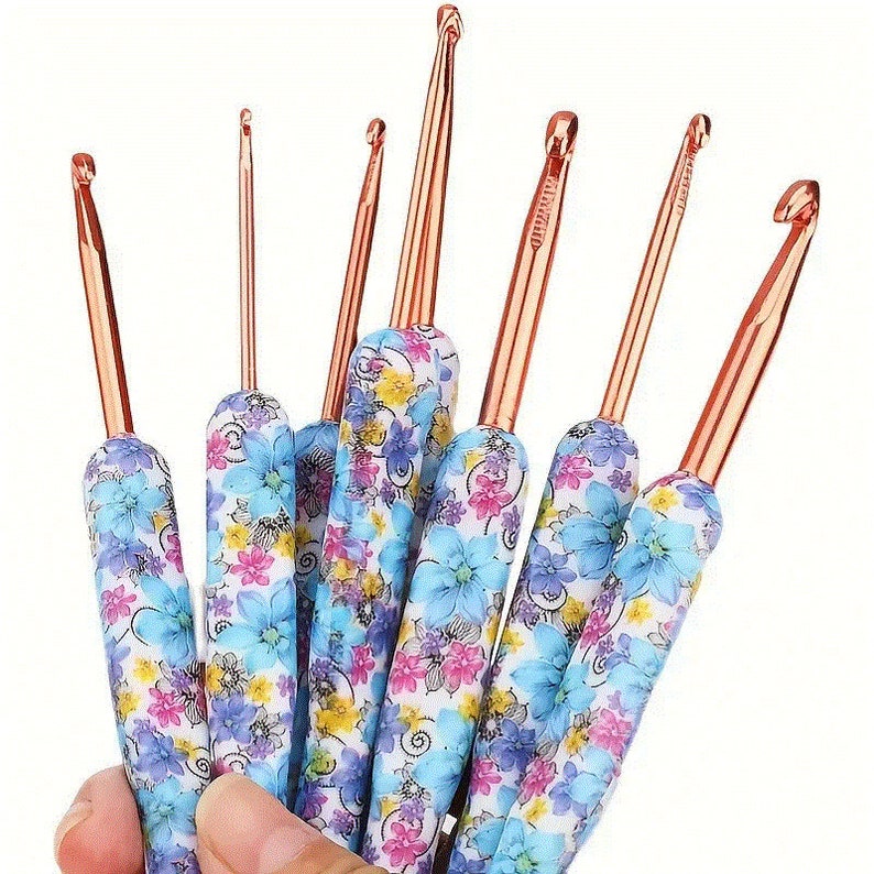 Pu&ograve; includere: Un set di uncinetti con steli in metallo color rame e manici con motivi floreali blu, rosa e bianchi. Gli uncinetti sono tenuti in una mano, mostrando diverse dimensioni e stili.
