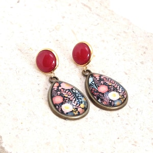 May include: A pair of drop earrings featuring a round, solid red stud top connected to a teardrop-shaped pendant. The pendant has a dark background with a colorful floral pattern of pink, white, and orange flowers, set in a bronze-toned metal frame.