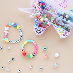 Könnte beinhalten: Ein Bastelset mit einem einhornförmigen Behälter voller bunter Perlen, Charms und Schnur. Zwei handgefertigte Armbänder werden gezeigt, eines mit der Aufschrift "MARGARITA" und das andere mit einem Regenbogenverlauf. Zusätzliche Perlen und Buchstabenperlen sind verstreut.