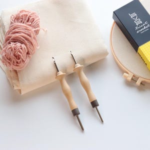 May include: Two punch needles with light wood handles and metal tips, a ball of dusty pink yarn, a folded piece of cream-colored fabric, and a wooden embroidery hoop with a black box labeled "Lavor tricot crochet PORTUGAL" on top.