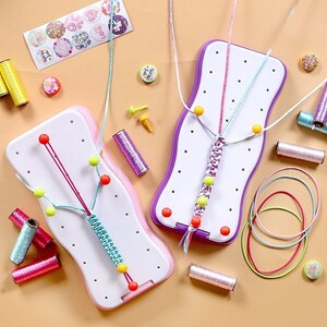 Può includere: Due tavole bianche e viola per creare braccialetti con filo colorato, perline e rocchetti di filo. Le tavole hanno una forma curva e sono circondate da materiali per l'artigianato, tra cui adesivi e spille.