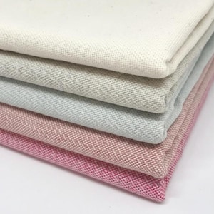 Op de afbeelding: Een stapel gevouwen stoffen stalen in verschillende kleuren. De bovenste laag is gebroken wit, gevolgd door grijs, lichtblauw en roze tinten. De stof heeft een getextureerde weefsel.