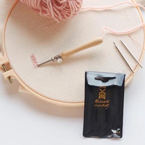 May include: An embroidery hoop with light beige fabric, a punch needle tool, and pink yarn. A black package with gold text reading "tricot crochet" is visible, alongside two needles and a ball of pink yarn, perfect for crafting.