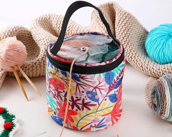 May include: A round yarn storage bag with a black handle and zipper. The bag features a colorful floral pattern with red, blue, and yellow leaves. A clear top allows visibility of the yarn inside. Knitting supplies and yarn balls are also visible.