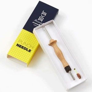 Puede incluir: Una herramienta de aguja de perforación con mango de madera y aguja de metal, presentada en una caja blanca. La caja está parcialmente abierta, revelando la herramienta y un pequeño accesorio. El embalaje es amarillo y azul marino, con las palabras "PUNCH NEEDLE".