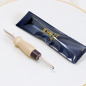 Könnte beinhalten: Ein Punch-Needle-Werkzeug mit einem hellbraunen Holzgriff und einem dunkelbraunen Sockel, neben einem marineblauen Etui mit goldenem Text. Das Werkzeug ist für Stickerei und Teppichhaken bestimmt. Die Artikel befinden sich auf einem weißen Stoff.