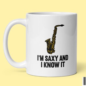 Puede incluir: Taza de cerámica blanca con un gráfico de saxofón negro y dorado y el texto "I'm saxy and I know it".