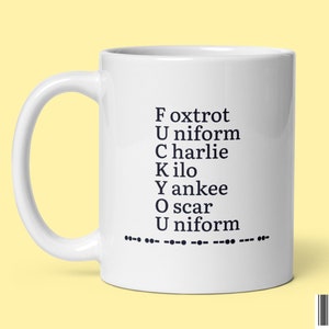 May include: White ceramic coffee mug with black text that reads "Fox Trot Uniform Charlie Kilo Yankee Oscar Uniform" and a row of dashes below the text.
