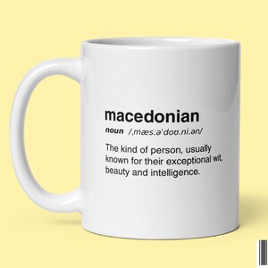 Puede incluir: Taza de cerámica blanca con la palabra "macedonian" impresa en negro, junto con una definición. La definición describe a una persona macedonia. La taza tiene un asa curvada.