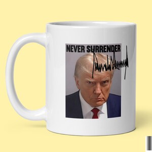 May include: White ceramic coffee mug with a black and white image of Donald Trump and the text "Never Surrender" with his signature underneath.